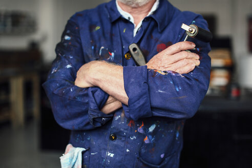 Älterer Handwerker mit verschränkten Armen in einer Druckwerkstatt, er hält eine Farbwalze und trägt einen mit Farbe bespritzten Overall, Mittelteil - CUF21077