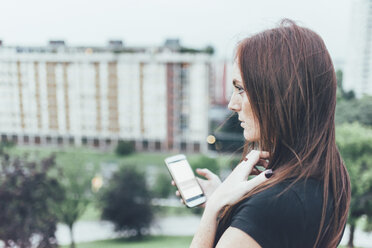 Young woman with long red hair holding smartphone and gazing above city - CUF21119