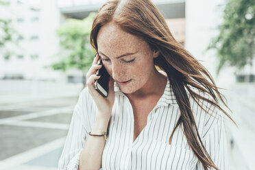 Junge Frau mit Sommersprossen und langen roten Haaren telefoniert mit ihrem Smartphone vor einem Bürogebäude - CUF21129