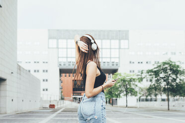 Junge Frau hört Kopfhörer mit der Hand auf dem Kopf vor einem Bürogebäude - CUF21131
