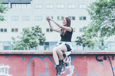 Junge Frau sitzt auf einer Graffiti-Wand und macht ein Selfie mit ihrem Smartphone - CUF21207