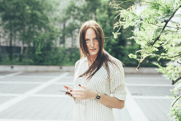 Portrait of young woman with freckles and long red hair holding smartphone in parking lot - CUF21212