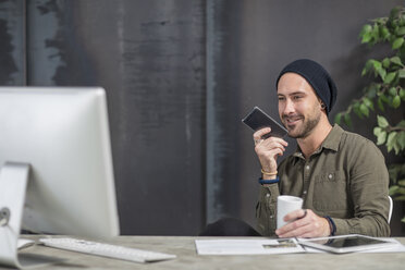 Lächelnder junger Mann mit Mobiltelefon am Schreibtisch im Büro - ZEF15525