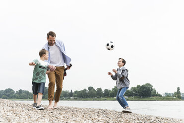 Glücklicher Vater mit zwei Söhnen und Fußball beim Spaziergang am Flussufer - UUF13915