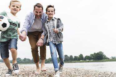 Glücklicher Vater mit zwei Söhnen und Fußball am Flussufer - UUF13917