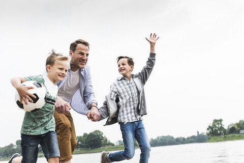 Glücklicher Vater mit zwei Söhnen und Fußball am Flussufer, lizenzfreies Stockfoto