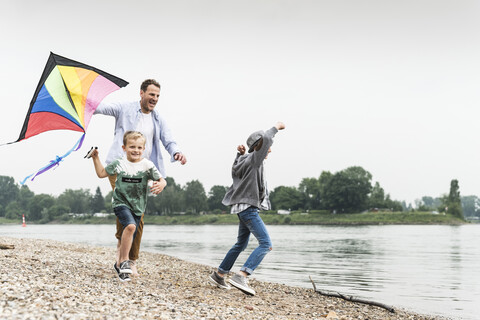 Glücklicher Vater mit zwei Söhnen beim Drachensteigenlassen am Flussufer, lizenzfreies Stockfoto
