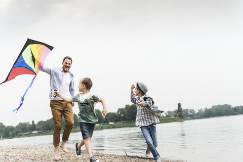 Glücklicher Vater mit zwei Söhnen beim Drachensteigenlassen am Flussufer, lizenzfreies Stockfoto