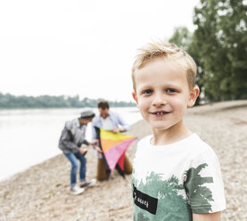Porträt eines lächelnden Jungen mit Vater und Bruder, der einen Drachen am Flussufer hält - UUF13934