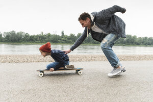 Glücklicher Vater schiebt seinen Sohn auf dem Skateboard am Flussufer - UUF13938