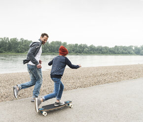 Glücklicher Vater läuft neben seinem Sohn auf dem Skateboard am Flussufer - UUF13939