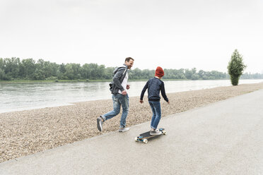 Glücklicher Vater läuft neben seinem Sohn auf dem Skateboard am Flussufer - UUF13940