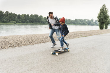 Glücklicher Vater läuft neben seinem Sohn auf dem Skateboard am Flussufer - UUF13941