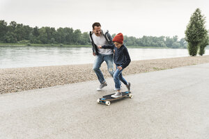 Glücklicher Vater läuft neben seinem Sohn auf dem Skateboard am Flussufer - UUF13941