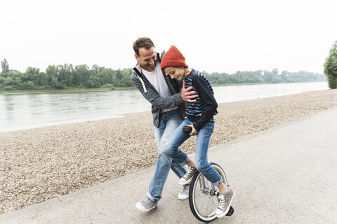 Glücklicher Vater unterstützt seinen Sohn auf dem Einrad am Flussufer, lizenzfreies Stockfoto