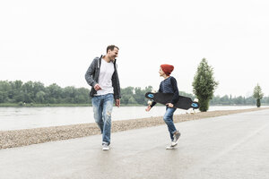 Glücklicher Vater und Sohn mit Skateboard am Flussufer - UUF13944