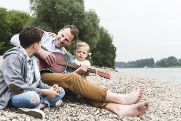 Happy father with two sons sitting at the riverside playing guitar - UUF13955