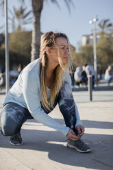 Spain, Barcelona, woman tying running shoe on beach promenade - MAUF01422