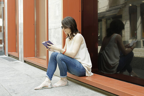 Frau sitzt vor einem Geschäft und liest in einem Notizbuch, Mailand, Italien - CUF21243
