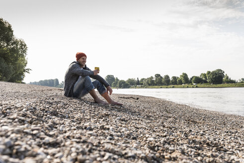 Mature man wearing red beanie and drinking coffee at Rhine riverbank - UUF13957
