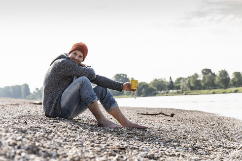 Mature man wearing red beanie and drinking coffee at Rhine riverbank - UUF13958