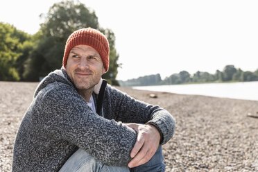 Mature man wearing red beanie sitting at Rhine riverbank - UUF13959