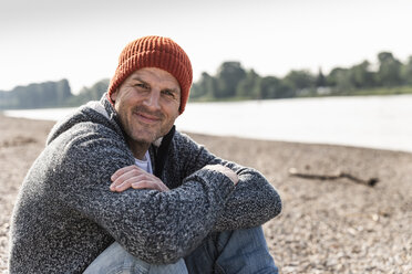 Mature man wearing red beanie sitting at Rhine riverbank - UUF13962