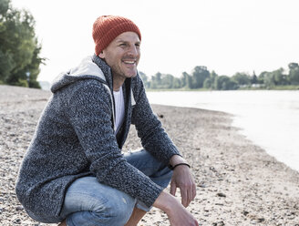 Smiling mature man with red beanie at Rhine riverbank - UUF13965