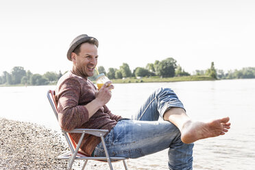 Mature man sitting on chair at Rhine riverbank - UUF13968