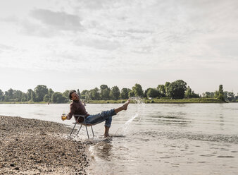 Mature man sitting on chair at Rhine riverbank - UUF13970