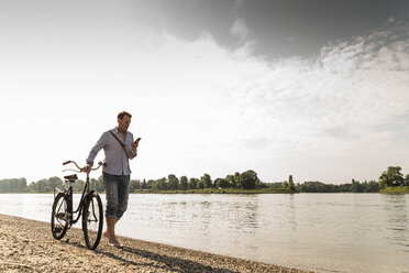 Älterer Mann mit Fahrrad und Smartphone am Rheinufer - UUF13973