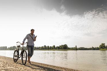 Mature man with bike using smartphone at Rhine riverbank - UUF13973
