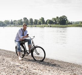 Älterer Mann mit Fahrrad und Smartphone am Rheinufer - UUF13978