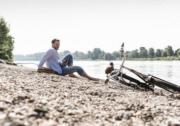 Mature man with bike and smartphone sitting at Rhine riverbank - UUF13979
