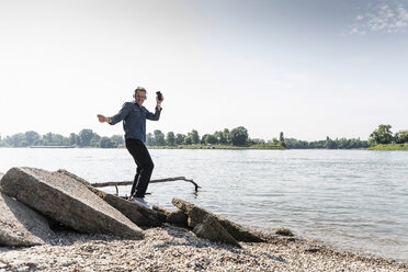 Mature man with smartphone dancing at Rhine riverbank - UUF13990