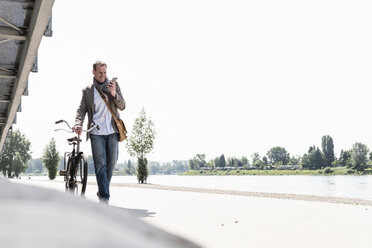Mature man with bike using smartphone at Rhine riverbank - UUF14000