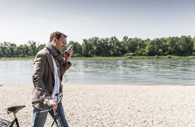 Mature man with bike using smartphone at Rhine riverbank - UUF14002