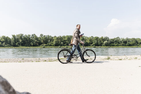 Mature man with bike using smartphone at Rhine riverbank - UUF14004