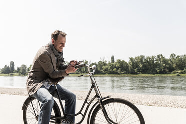 Mature man with bike using smartphone at Rhine riverbank - UUF14009