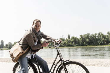 Mature man with bike using smartphone at Rhine riverbank - UUF14010