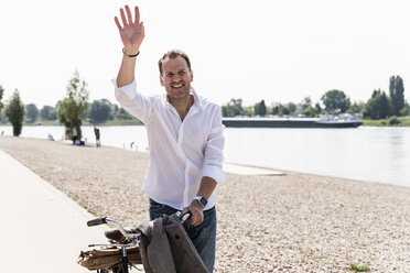 Mature man with bike waving at Rhine riverbank - UUF14013