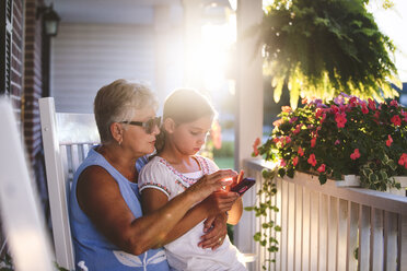 Mädchen und Großmutter spielen Smartphone-Spiel auf Veranda bei Sonnenuntergang - ISF07684