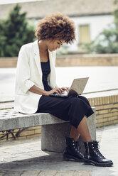 Modische junge Frau mit lockigem Haar sitzt auf einer Bank und benutzt einen Laptop - JSMF00214