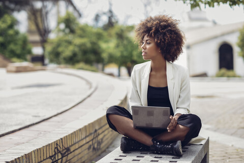 Modische junge Frau mit lockigem Haar sitzt auf Bank mit Laptop, lizenzfreies Stockfoto