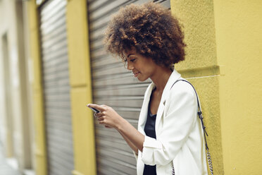 Lächelnde junge Frau mit lockigem Haar, die auf ihr Mobiltelefon schaut - JSMF00227