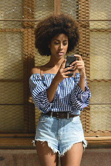 Porträt einer modischen jungen Frau mit lockigem Haar, die auf ihr Smartphone schaut - JSMF00253
