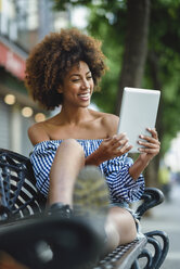 Porträt einer lachenden jungen Frau, die auf einer Bank sitzt und ein Tablet benutzt - JSMF00265