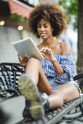 Porträt einer jungen Frau mit lockigem Haar, die auf einer Bank sitzt und ein Tablet benutzt - JSMF00267