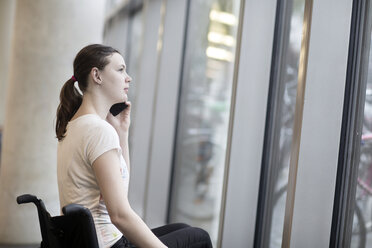 Young woman using wheelchair gazing through entrance window talking on smartphone - CUF21384