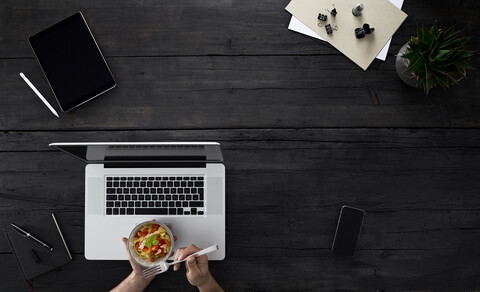 Person mit Laptop und Snack am Schreibtisch sitzend, lizenzfreies Stockfoto